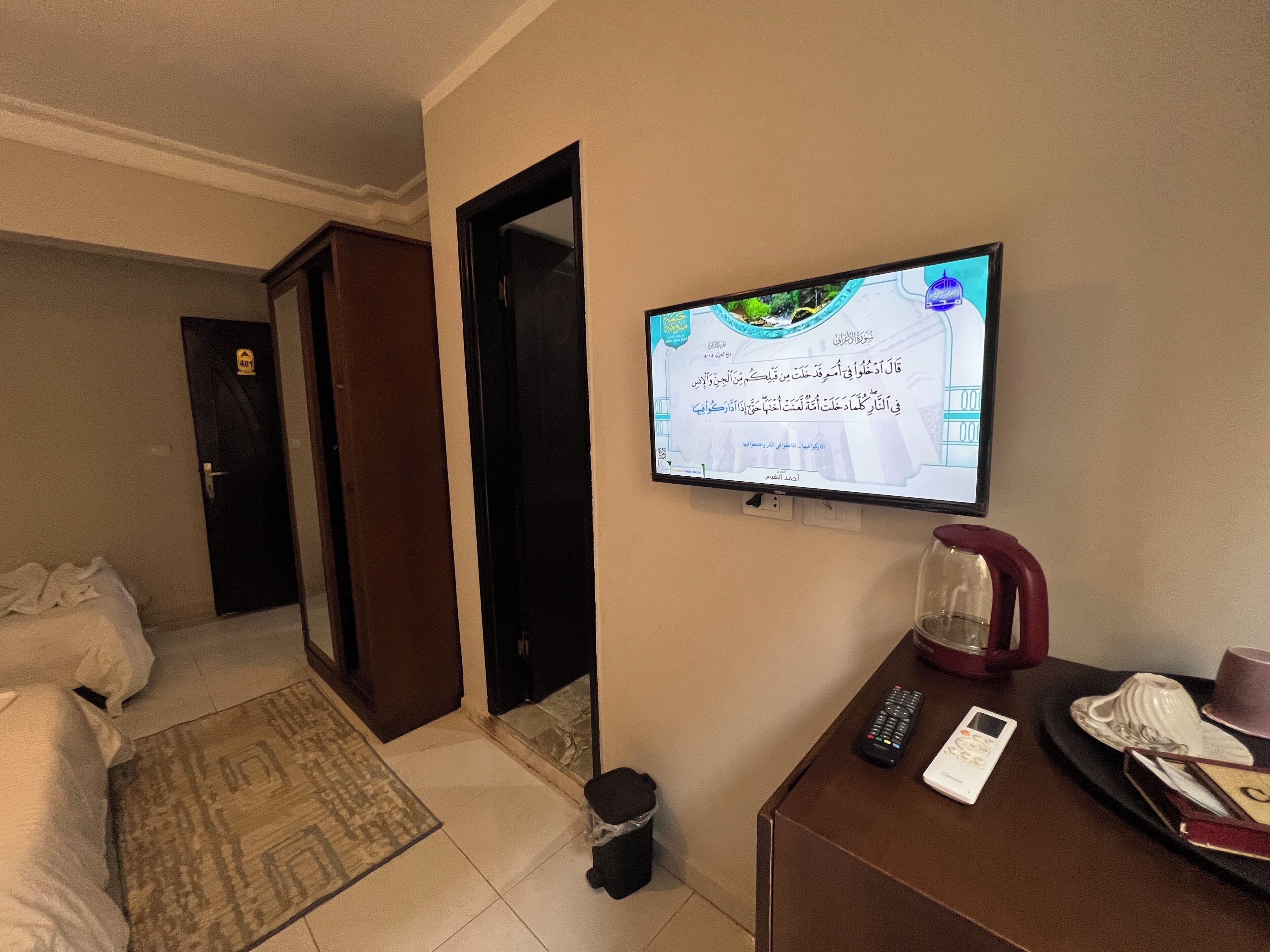 A hotel room with a television mounted on the wall displaying Arabic text, a bed with white linens, a small table with a remote control, a glass pitcher, and a tray with a cup and a hat.