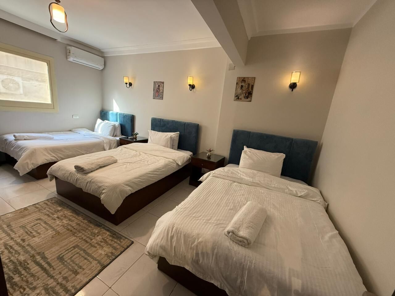 A hotel room with three beds, each with a blue headboard and white bedding. There are wall-mounted lights and a small rug on the floor.