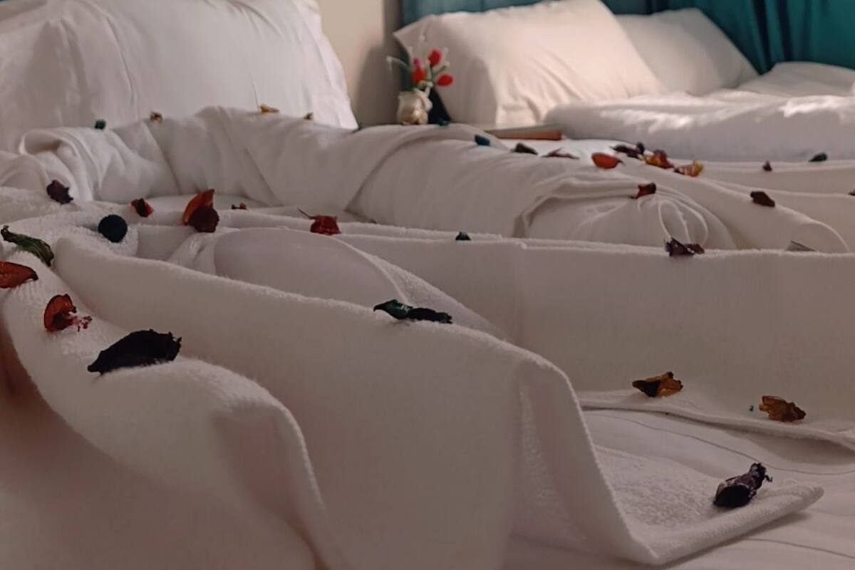 A hotel room with two beds, each with white bedding and a headboard. There is a window with teal curtains, and a small vase with flowers on the nightstand.