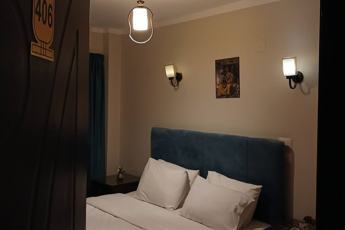 A hotel room with a bed, a nightstand, a wall-mounted light, and a painting.