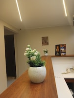 Una recepción con un escritorio de madera, un jarrón blanco con textura salpicada que contiene flores blancas, un cuadro en la pared y una pequeña planta en maceta.