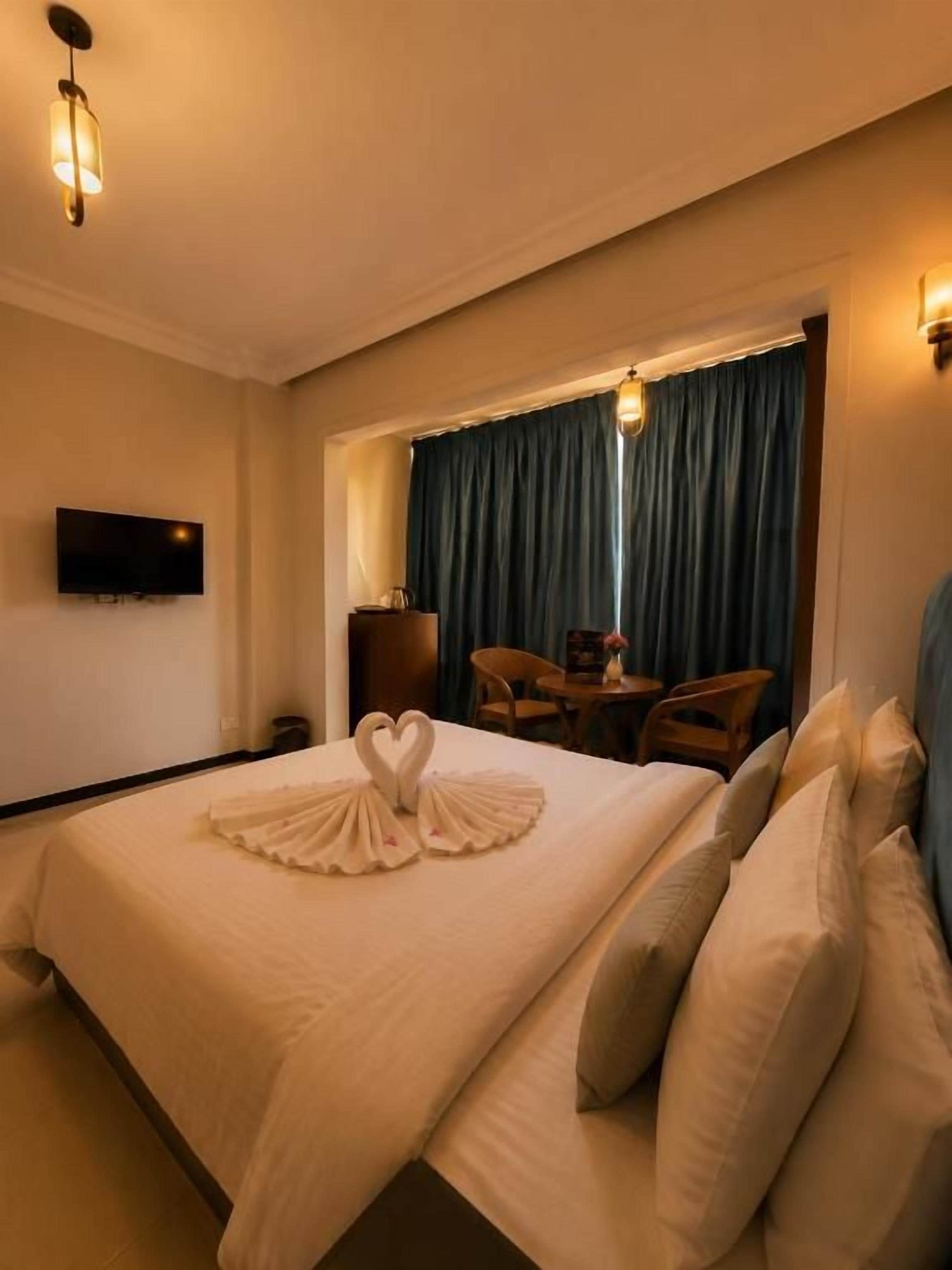 Ein Hotelzimmer mit einem großen Bett, einem flachbildfernseher an der Wand, einem Fenster mit Vorhängen und einem kleinen Tisch mit Stühlen.