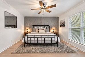 Een slaapkamer met een bed, nachtlampen, een plafondventilator, een groot raam met zonwering en een kamerkleed met dessin.