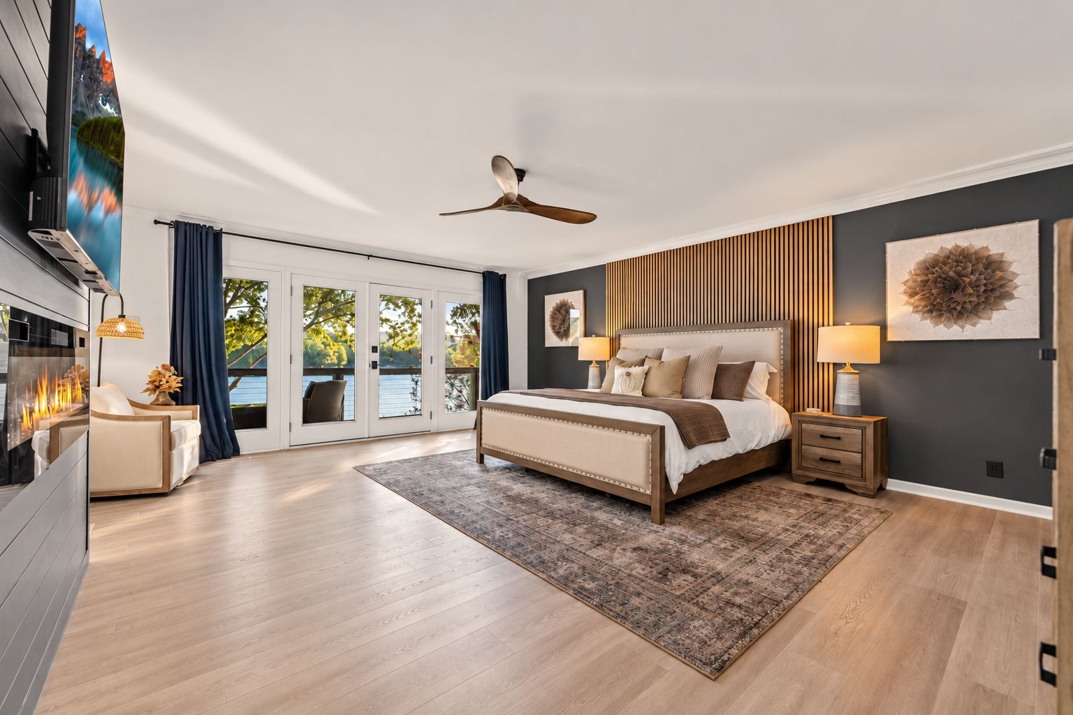 A modern bedroom with a large bed, a ceiling fan, a fireplace, and a view through the glass doors.