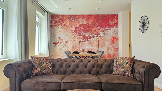 A modern living room with a tufted sofa, a wooden coffee table, and a dining area with a red textured wall. - Ruby - Right next to the castle, balcony, PS4 & Stream (Montabaur)