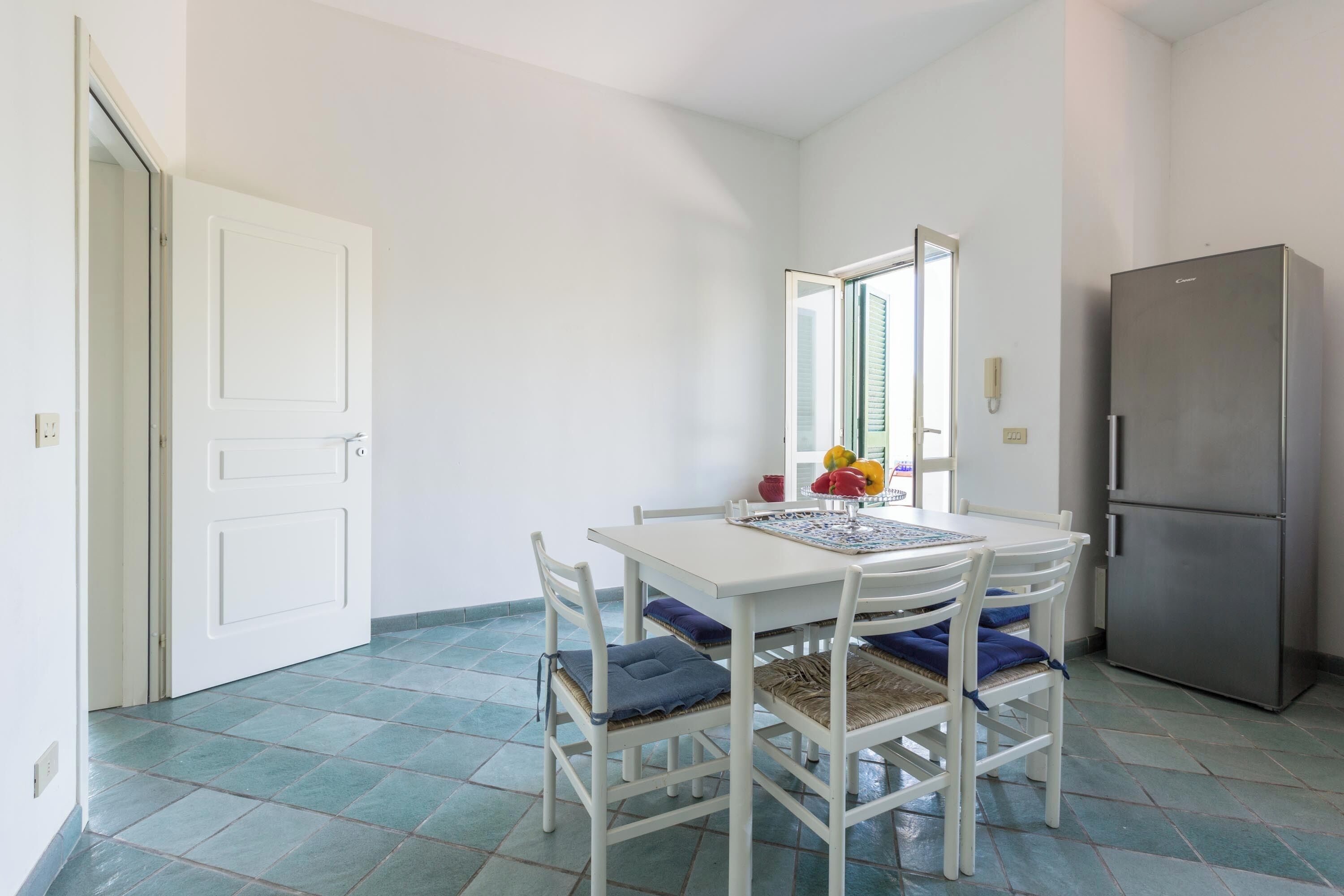 A small dining area with a white table, blue cushioned chairs, a stainless steel refrigerator, and a tiled floor.