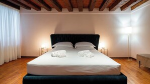 A hotel room with a large bed, wooden beams on the ceiling, two bedside tables with lamps, and a floor lamp. - San Beneto  (Venice)