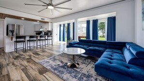 A modern living room with a blue sectional sofa, a round coffee table, and a kitchen area with a refrigerator and bar stools.