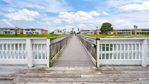 A wooden boardwalk leading to a building complex with multiple units, a clear sky, and a palm tree.