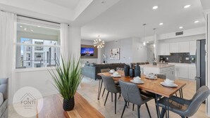 Una cocina moderna con armarios blancos, un comedor con mesa y sillas de madera, y un amplio ventanal con cortinas.