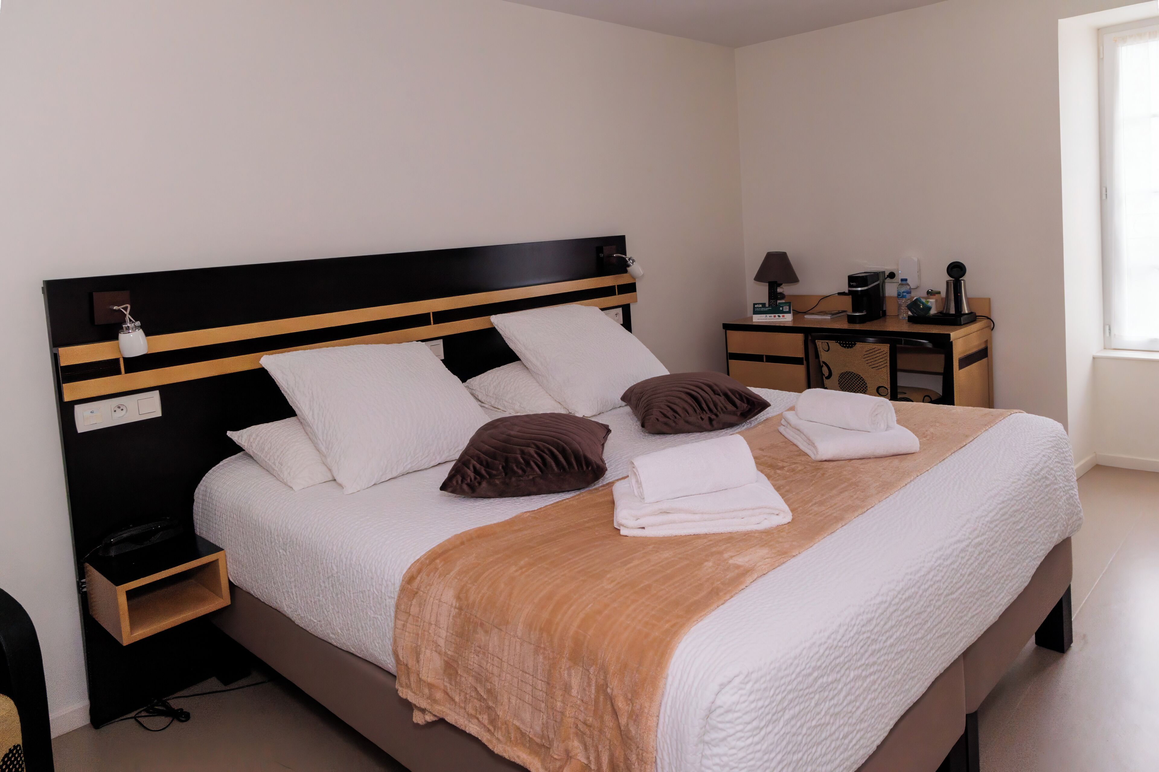 A neatly made bed with a brown headboard, white pillows, and a brown throw blanket. There is a small wooden nightstand with a lamp and a power outlet. A desk with a lamp, a coffee maker, and a small plant is visible in the corner.
