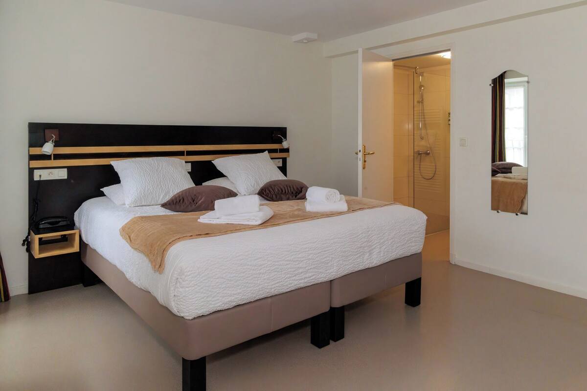 Een opgemaakt bed met wit en bruin beddengoed, een nachtkastje met een lamp, en een deur naar een douche.