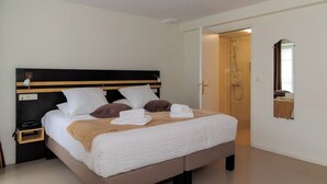 Una cama bien hecha con ropa de cama blanca y marrón, una mesita de noche con una lámpara y una puerta que da a una zona de ducha.