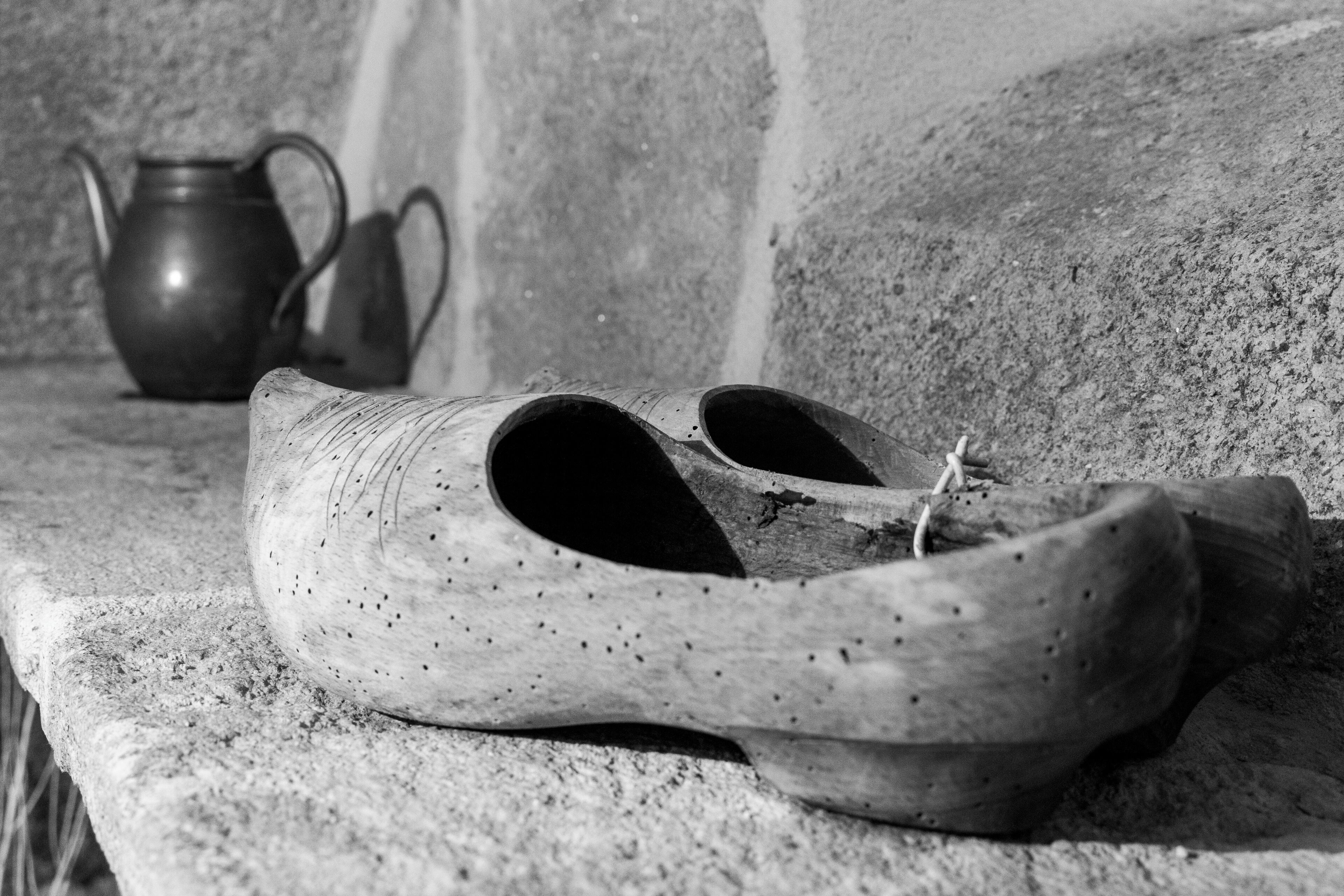 A pair of wooden shoes on a stone ledge with a metal pitcher and a metal jug in the background.