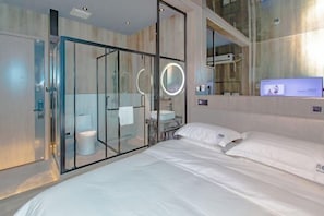 A modern hotel room with a glass-enclosed shower, a bed with white linens, and a built-in TV on the wall. - QIngGuo intelligence Hotel (Jiashan)
