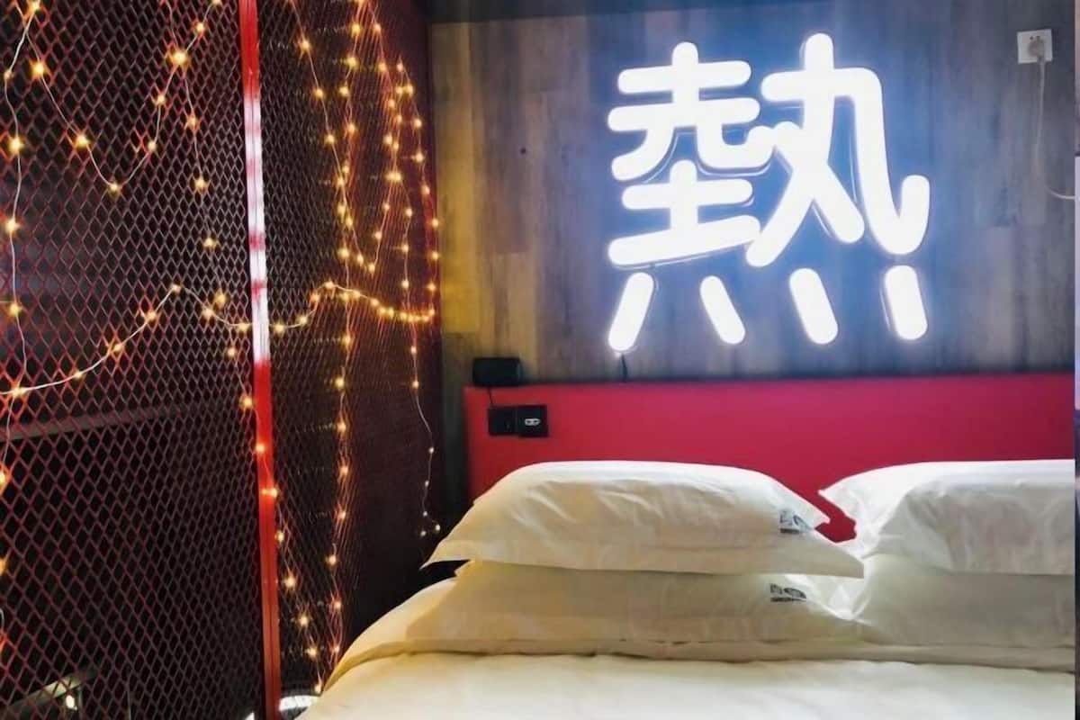 Camera d'albergo con un letto, cuscini bianchi, una testata rossa e un cartello luminoso con caratteri cinesi.