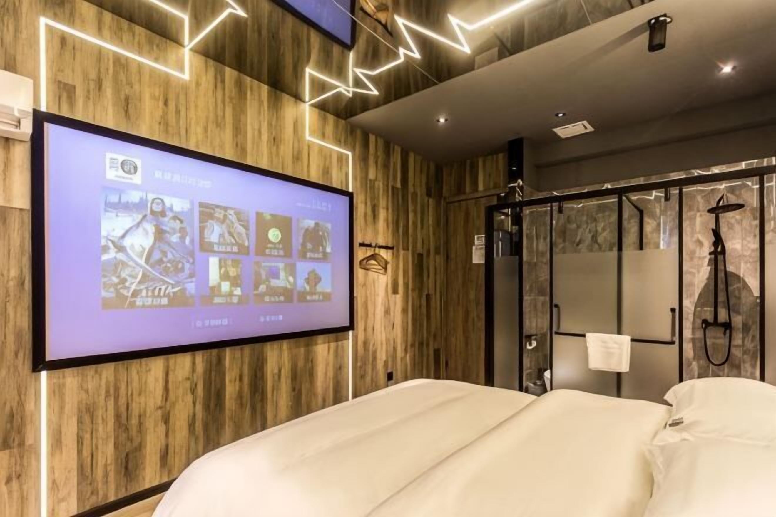 Une chambre d’hôtel moderne dotée d’un grand téléviseur fixé au mur, de lambris en bois, d’un espace douche et d’un lit avec une literie blanche.