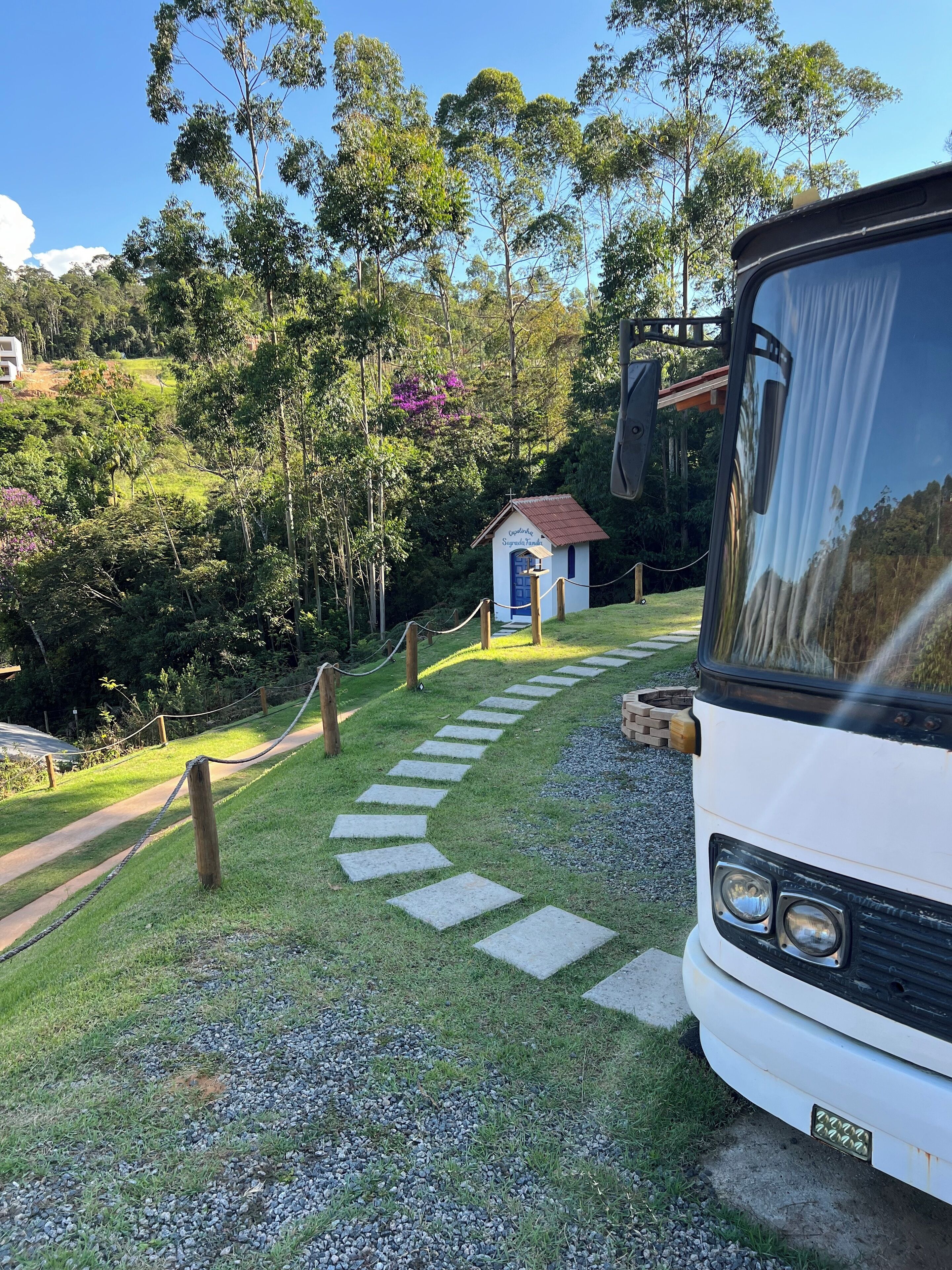 A white bus parked on a gravel path leading to a small building with a red roof and a blue door, surrounded by lush greenery and trees.