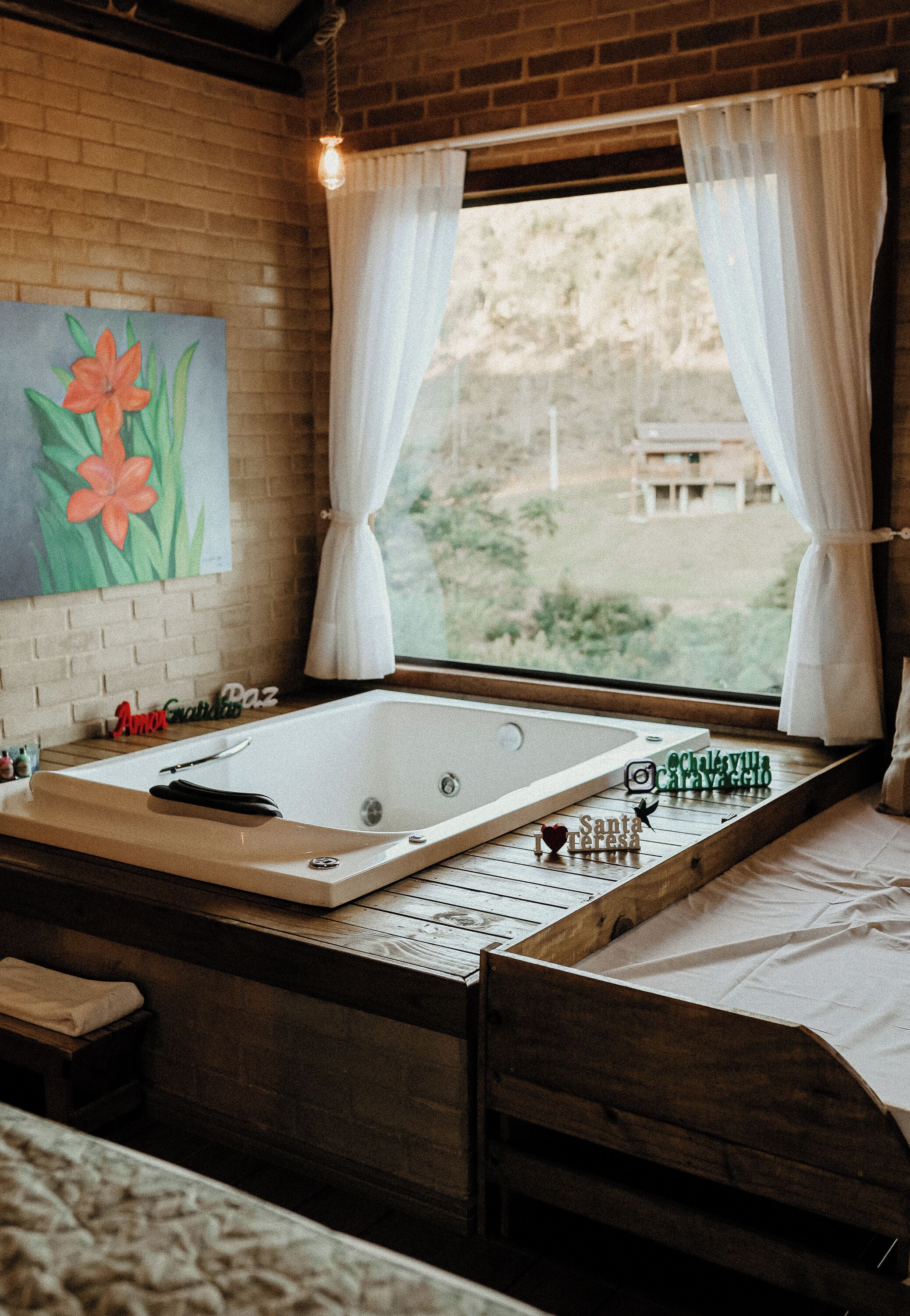 A room with a large bathtub, a wooden bench, a painting of flowers, and a view through a window with curtains.