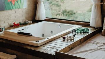 A room with a large bathtub, a wooden bench, a painting of flowers, and a view through a window with curtains.