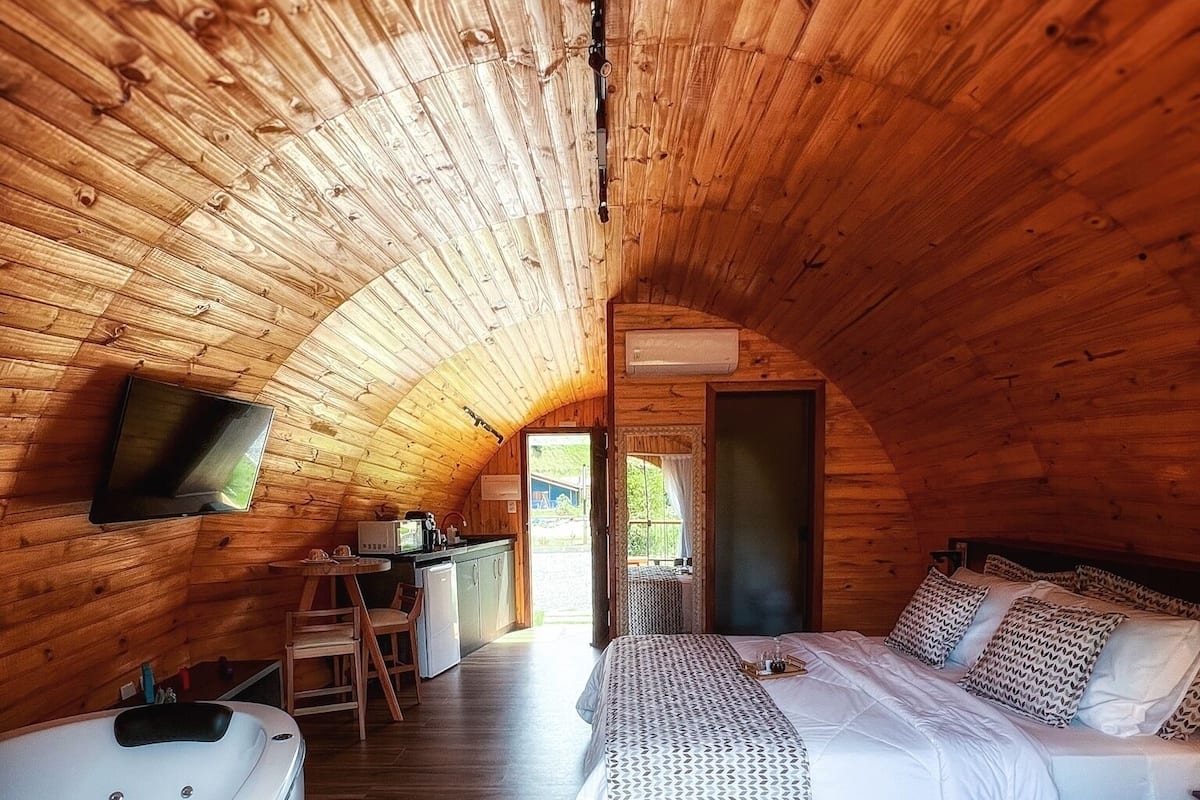 A cozy, wooden cabin bedroom with a bed, a small desk, a bathtub, and a TV mounted on the wall.
