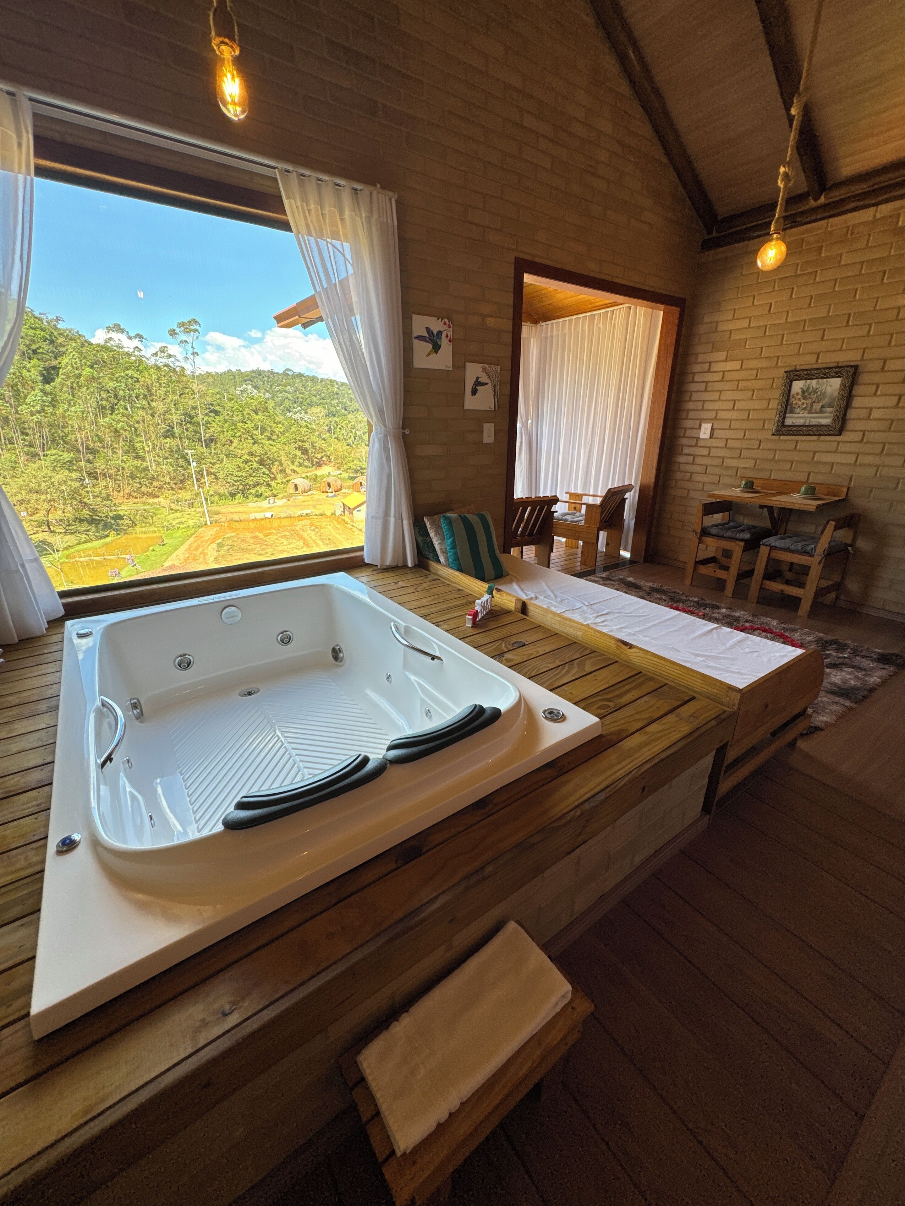 Una habitación con piso de madera, una bañera grande, un banco de madera, una mesa con sillas y vistas a un área boscosa a través de una ventana.