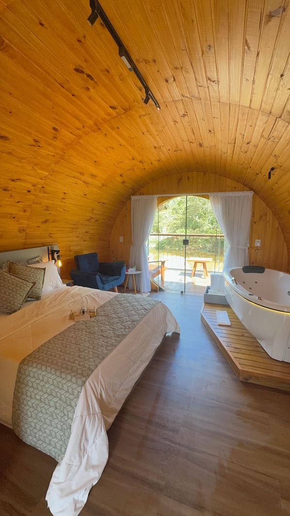 A cozy, wooden-roofed room with a large bed, a small table, a chair, and a bathtub. - Chalés Villa Caravaggio (Santa Teresa)