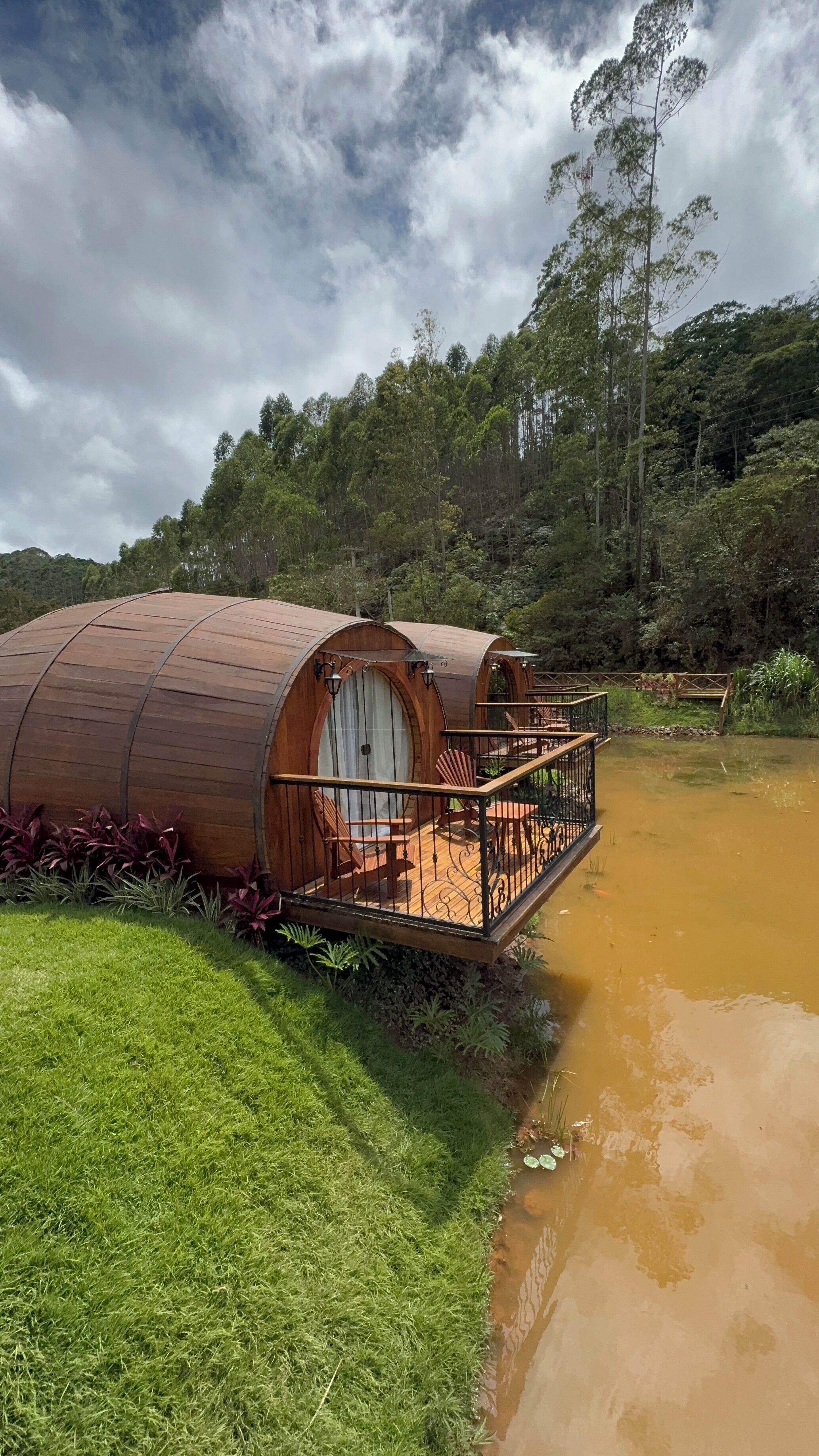Estructura cilíndrica de madera con balcón, ubicada junto a una masa de agua, rodeada de exuberante vegetación y árboles.