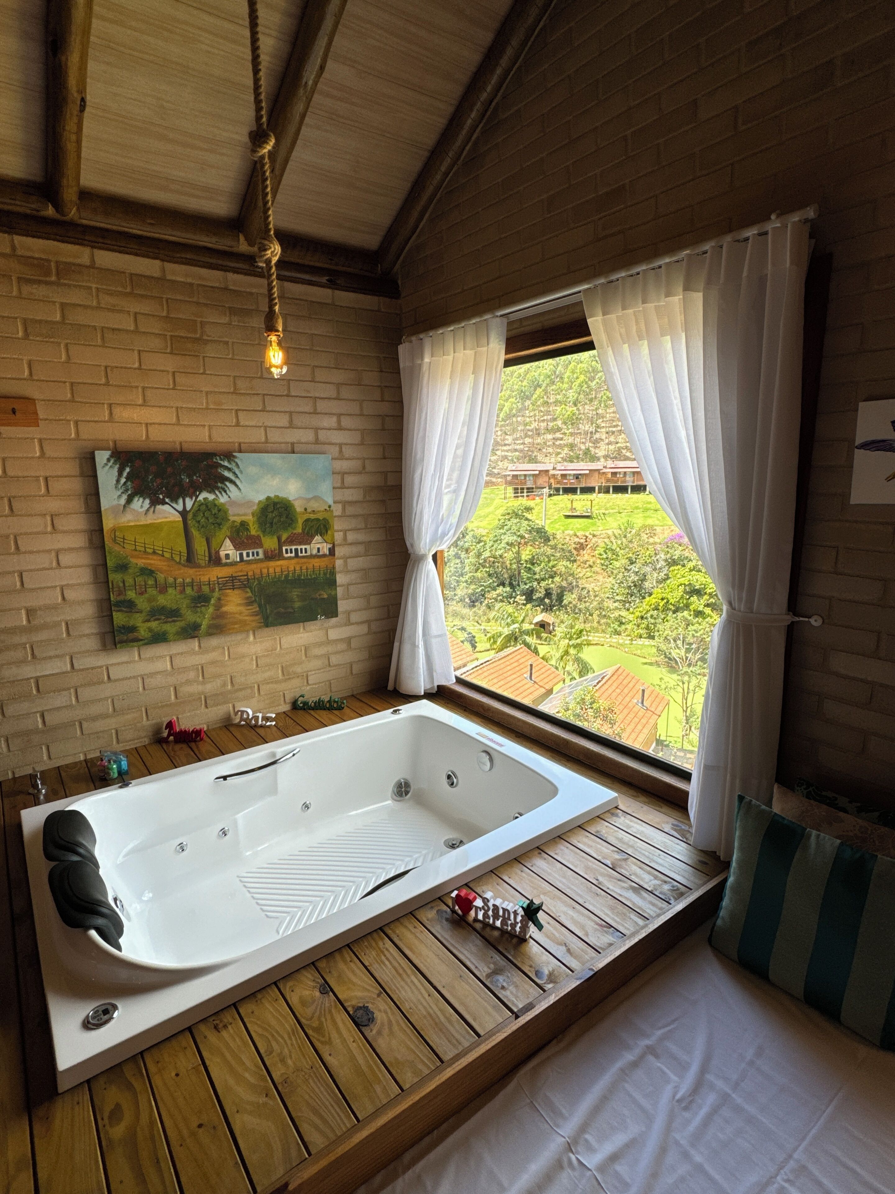 Una habitación con piso de madera, una bañera grande, un cuadro en la pared y vista a un paisaje verde a través de una ventana con cortinas blancas.