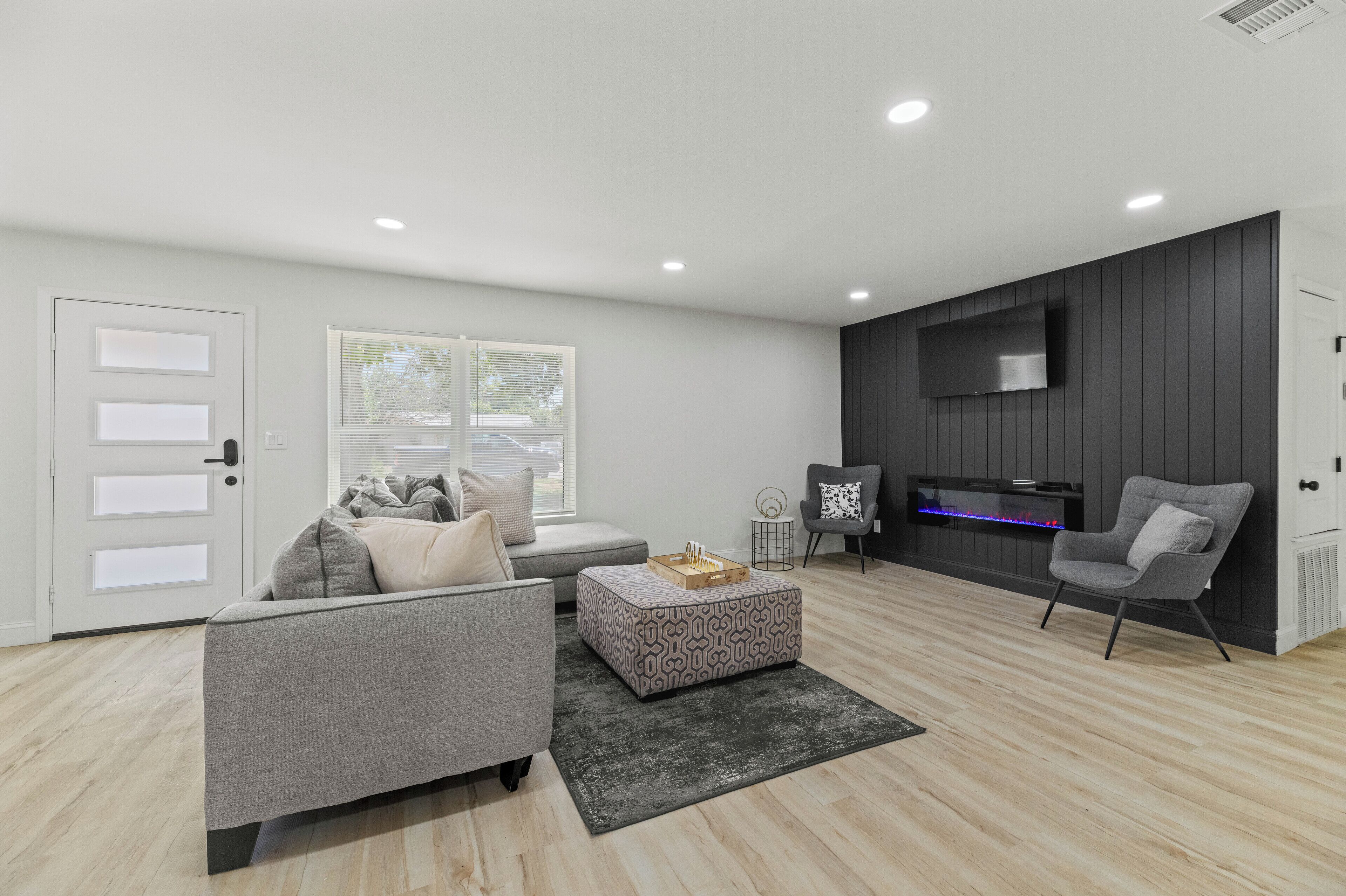 A modern living room with a grey sofa, a patterned ottoman, and a flat-screen TV mounted on a dark wall.