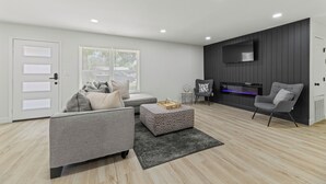 Un salon moderne avec un canapé gris, un pouf à motifs et un téléviseur à écran plat fixé sur un mur foncé.