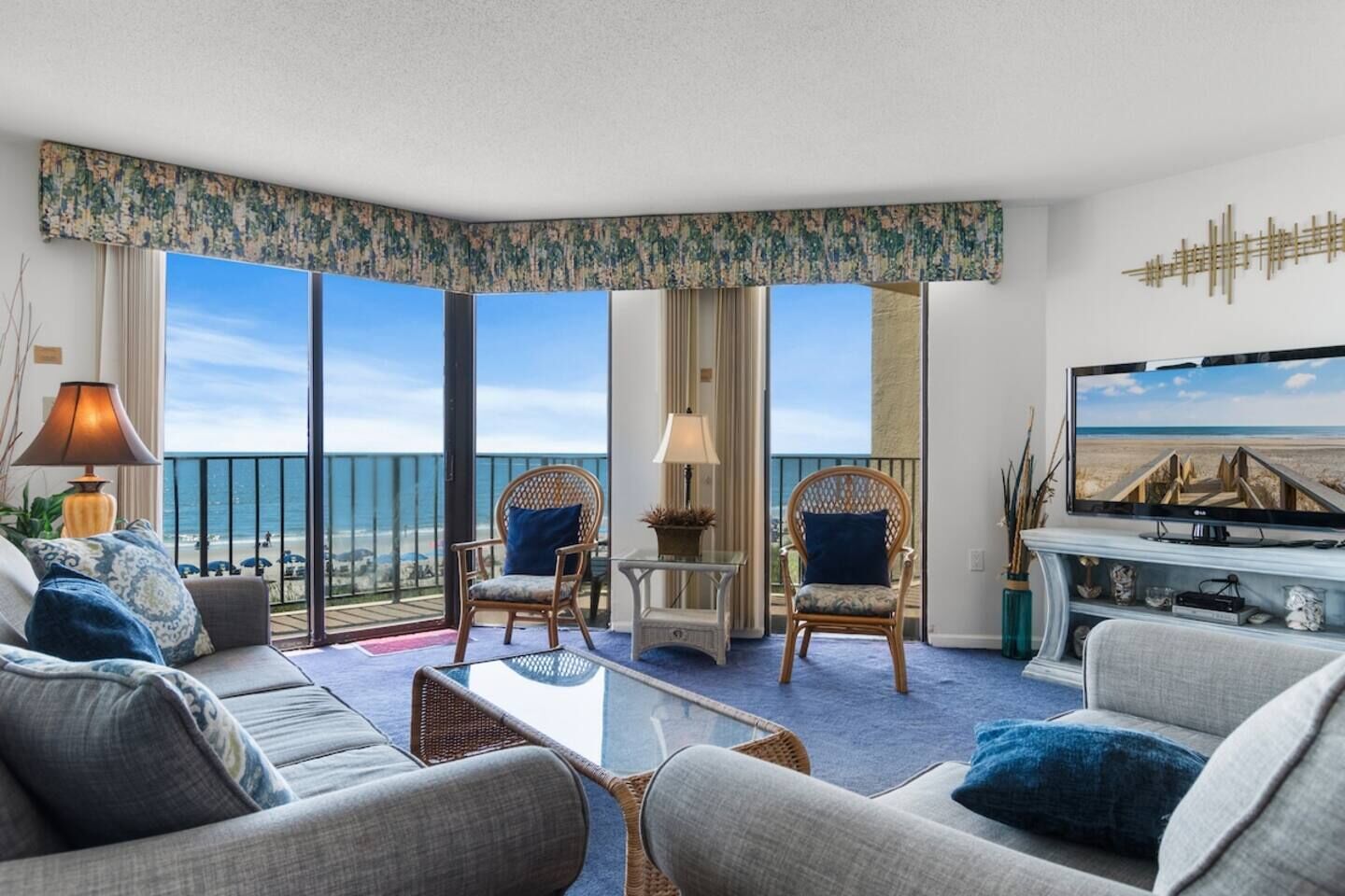 A hotel room with a balcony, two wicker chairs, a sofa, a coffee table, a TV, and a view of the beach.