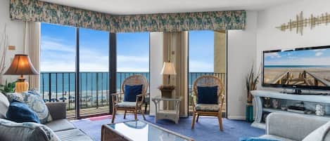 A hotel room with a balcony, two wicker chairs, a sofa, a coffee table, a TV, and a view of the beach.