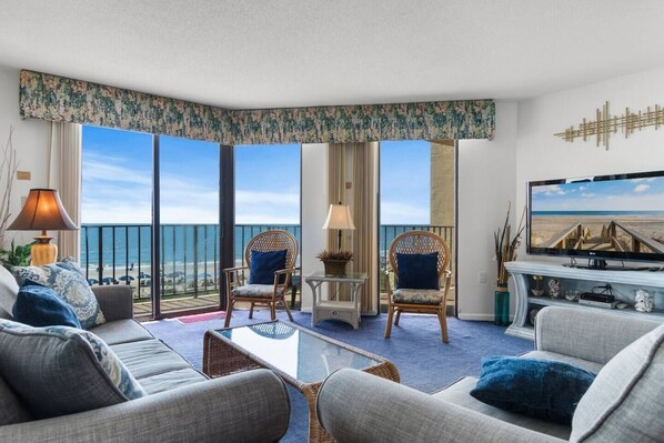 A hotel room with a balcony, two wicker chairs, a sofa, a coffee table, a TV, and a view of the beach.