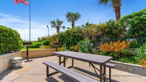 A picnic table with benches, surrounded by lush greenery and a palm tree, under a clear sky.