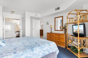 Un dormitorio con una cama, un tocador, un televisor sobre un soporte de madera y un baño visible a través de una puerta abierta.