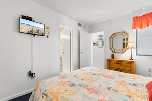 Quarto com cama, cômoda, televisão fixada na parede e janela com persianas.