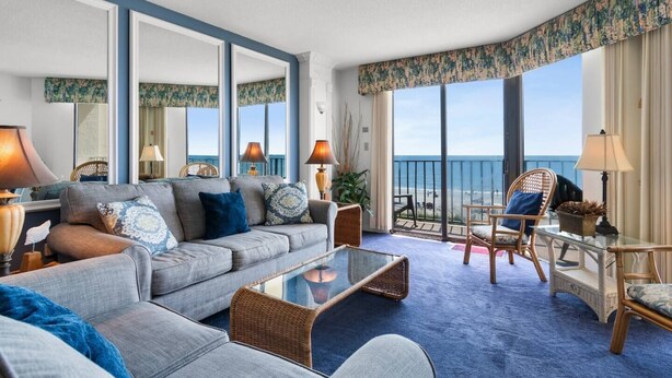 A hotel room with a large sofa, a glass coffee table, wicker chairs, a balcony with a view of the sea, and a patterned curtain.