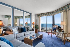 Habitación de hotel con un sofá grande, una mesa de centro de vidrio, sillas de mimbre, un balcón con vista al mar y una cortina estampada.