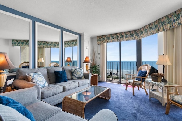 Habitación de hotel con un sofá grande, una mesa de centro de vidrio, sillas de mimbre, un balcón con vista al mar y una cortina estampada.