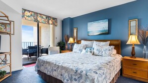 A bedroom with a large bed, a desk, and a chair, a balcony with a view, and a painting on the wall.