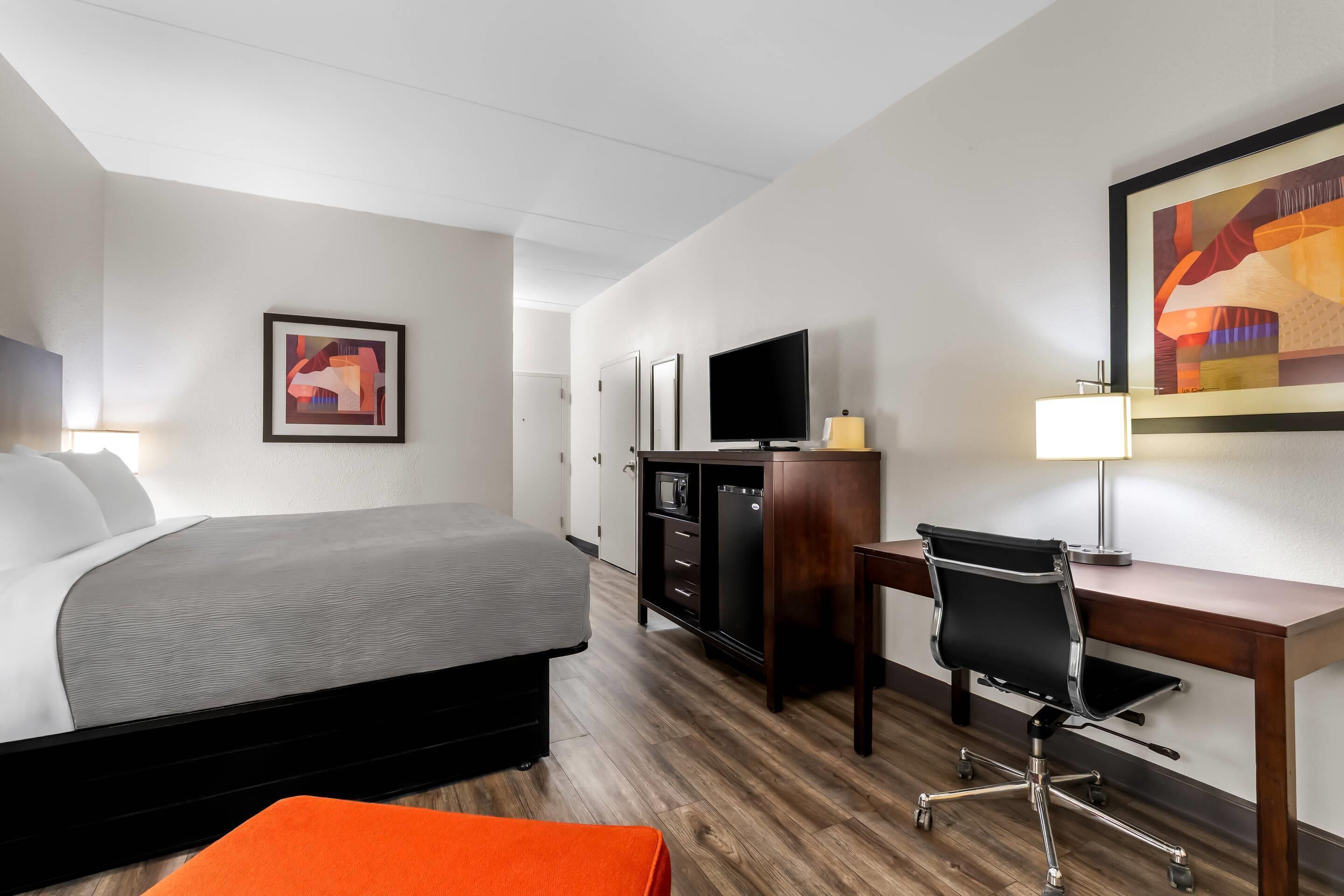 A hotel room with a bed, desk, chair, TV, and artwork on the walls.