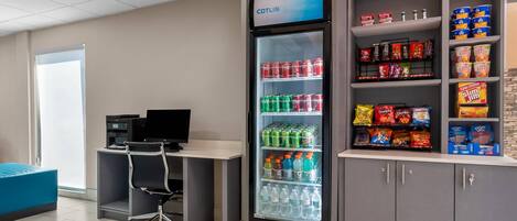 A vending machine with a variety of drinks and snacks, a desk with a computer, and a blue seating area.