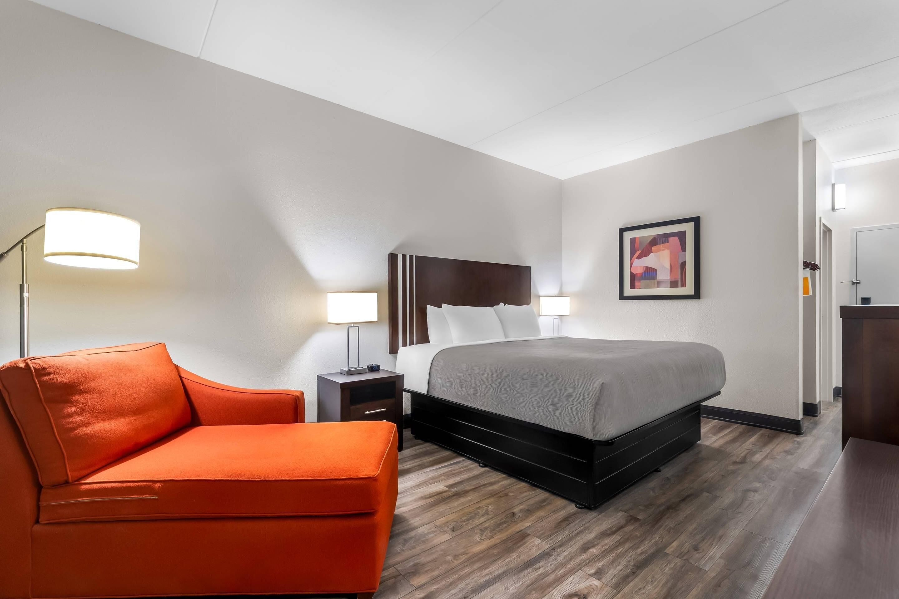 A hotel room with a bed, a nightstand, a lamp, a chair, and a painting on the wall.