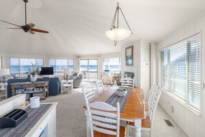 Ein geräumiger Wohnbereich mit Esstisch aus Holz, weißen Stühlen, Deckenventilator und großen Fenstern mit Meerblick.