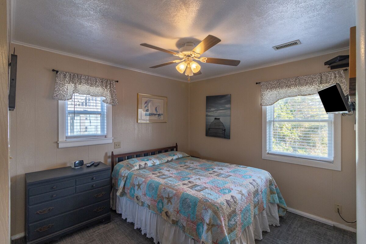 A bedroom with a bed, dresser, ceiling fan, two windows with curtains, and a mounted TV.