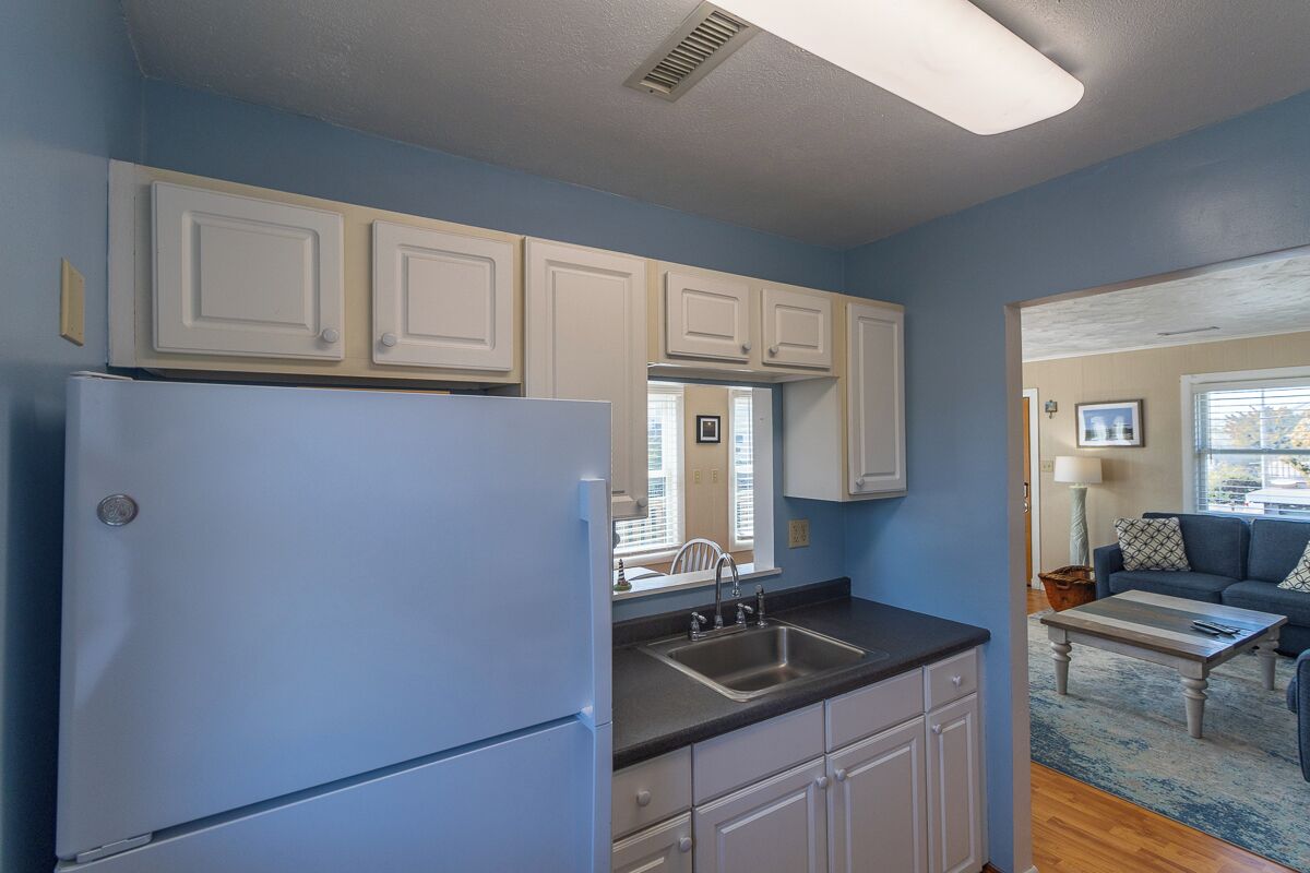 A kitchen with white cabinets, a refrigerator, and a sink. There is a window with blinds and a view of the outside.