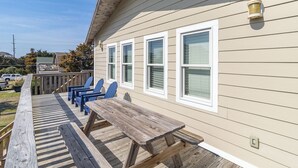 A wooden deck with a picnic table and blue chairs, attached to a beige house with white trim and multiple windows. - Pet-friendly oceanside Hatteras Island home by Sun Realty (Avon)