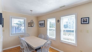 A dining area with a table and chairs, large windows with blinds, a ceiling light fixture, and a framed picture on the wall. - Pet-friendly oceanside Hatteras Island home by Sun Realty (Avon)