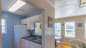 A compact kitchen with white cabinets and a refrigerator, a dining area with a table and chairs, and a living room with a patterned armchair. - Pet-friendly oceanside Hatteras Island home by Sun Realty (Avon)